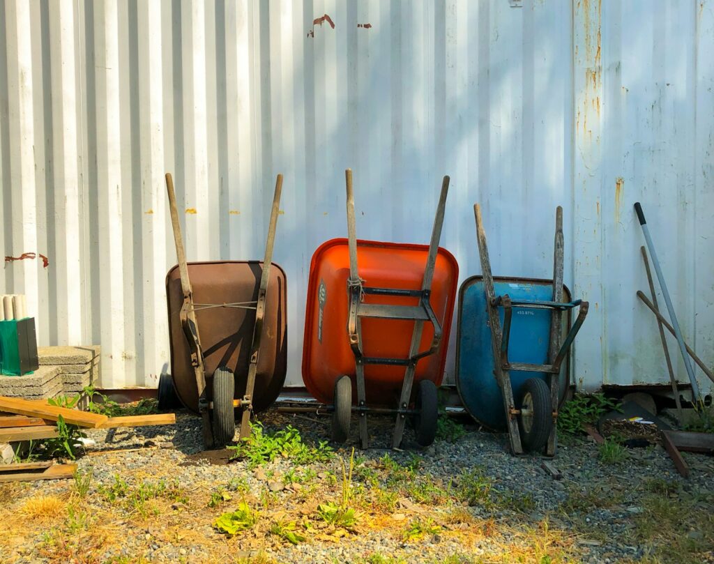 Three wheel barrows leaning up against a white fence sitting on top of a dirt and grass lawn.