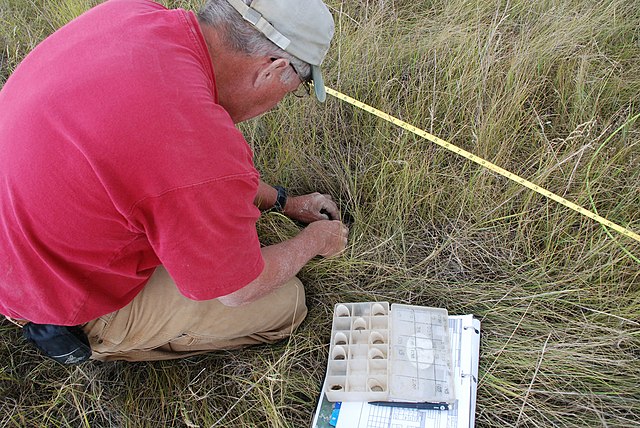 A person kneeling on grassy terrain, performing a soil test near a yellow measuring tape, with a soil testing kit and documentation open beside them.