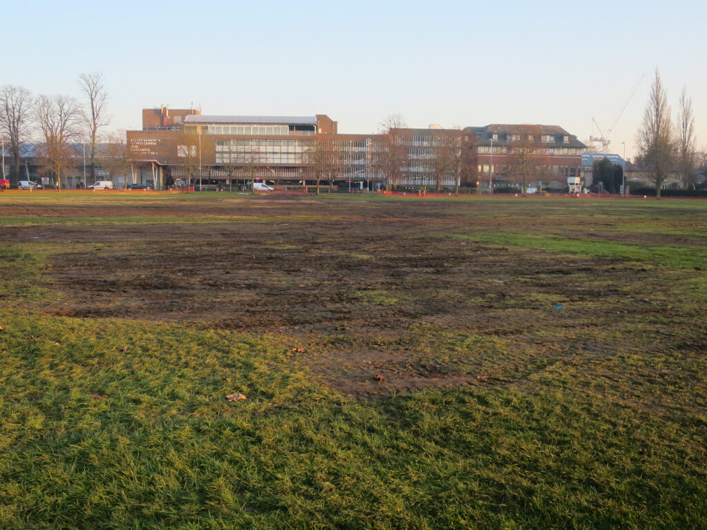 A field with patchy grass and large areas of bare soil, with a building in the background.